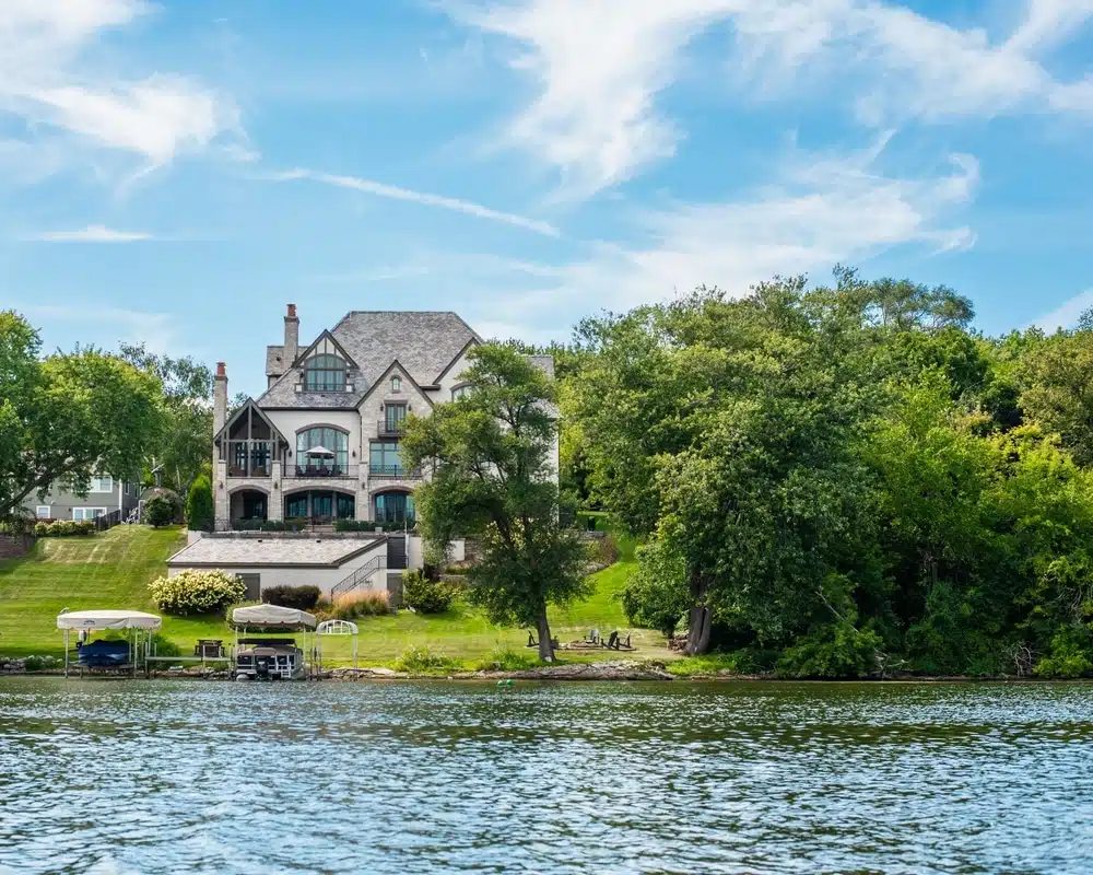 Lakeside mansion with stone and stucco exterior, lush lawn, and boats on a dock by a calm lake.