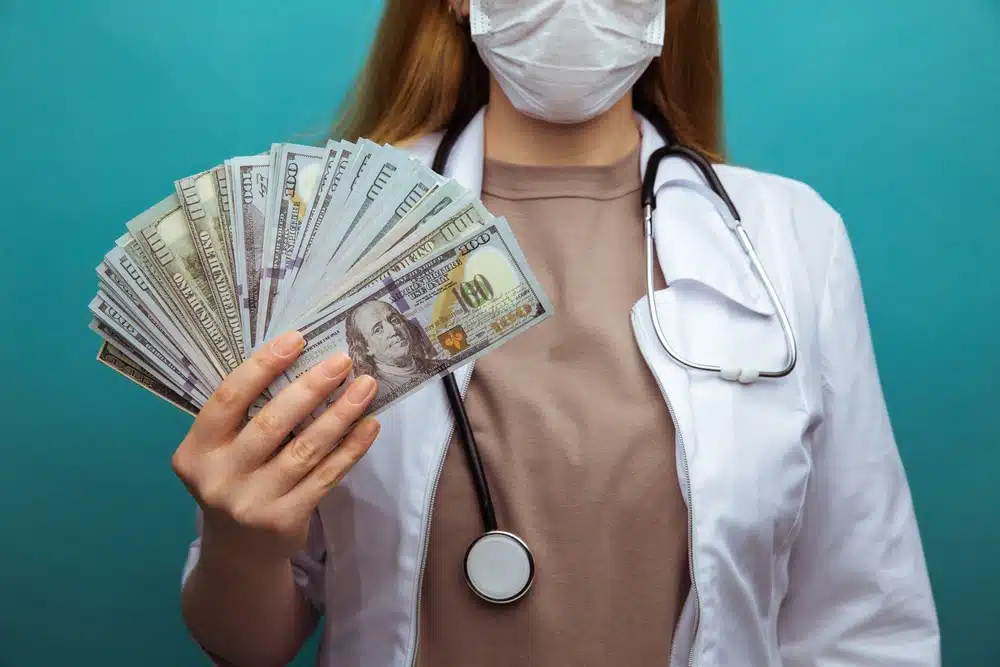 Female doctor in a mask and white coat holds a fan of us $100 bills, against a teal background.