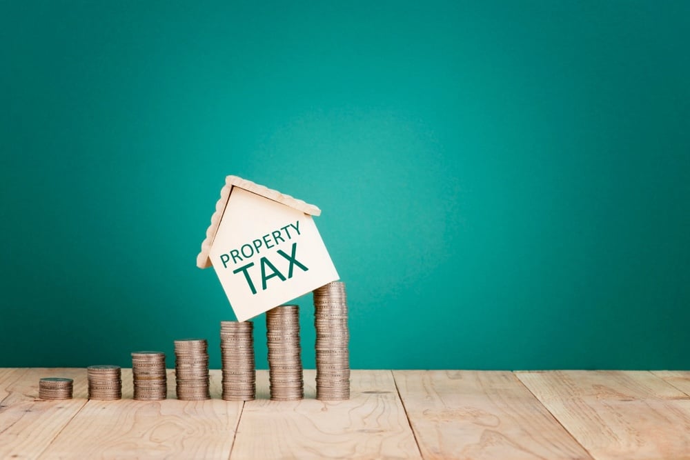 Small white house model labeled 'Property Tax' leaning on rising stacks of coins on a wooden surface with a teal background.