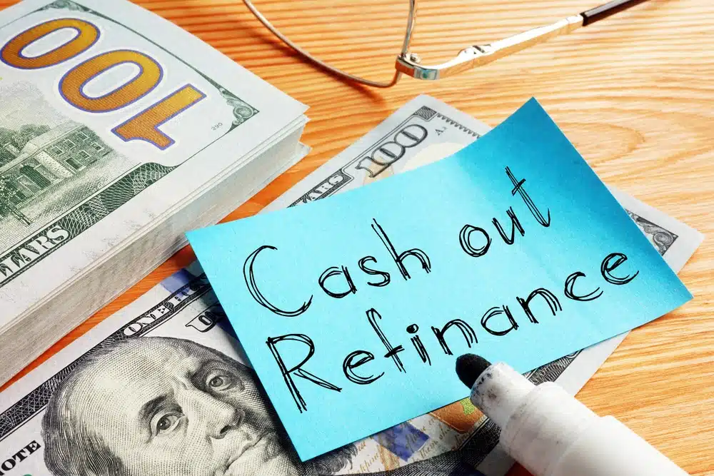 Blue sticky note with 'Cash out Refinance' on top of cash and a wooden desk, with glasses nearby.